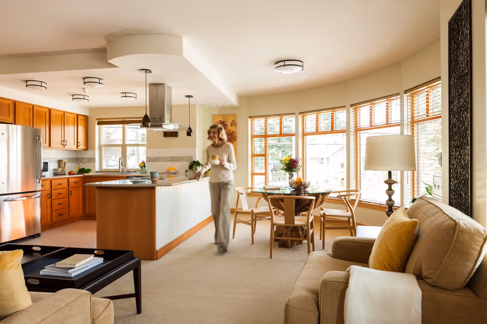 Open-plan interior showing a kitchen, dining area, and living room with a woman holding a mug walking through the space.