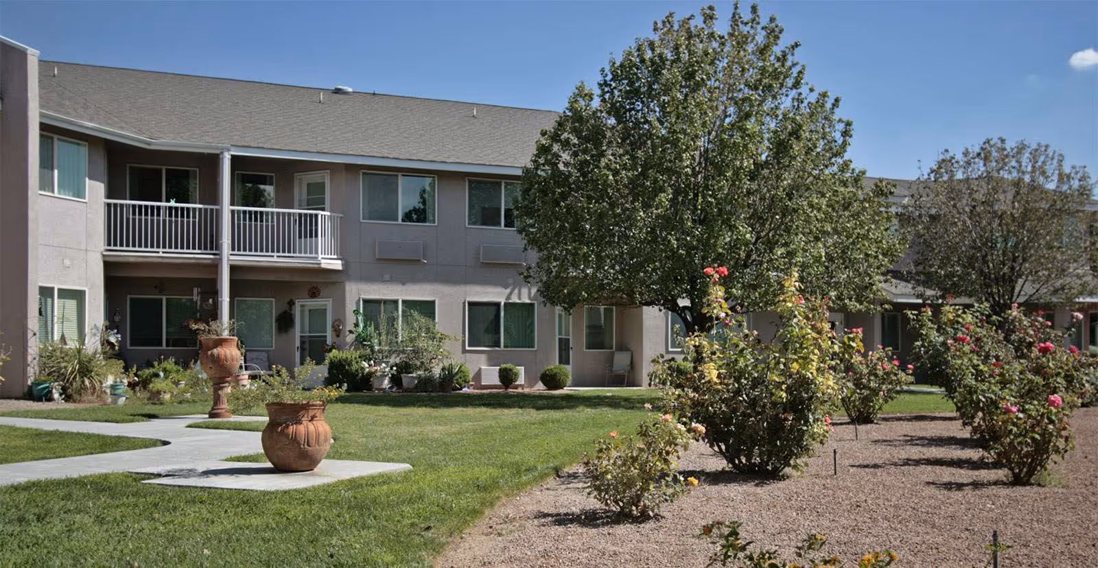Two-story residential building with balconies facing a manicured lawn, potted planters, trees, and flowering shrubs under a blue sky.