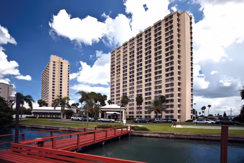 Tall residential buildings with multiple floors next to a body of water, featuring a red wooden bridge, palm trees, parked cars, and a partly cloudy sky.