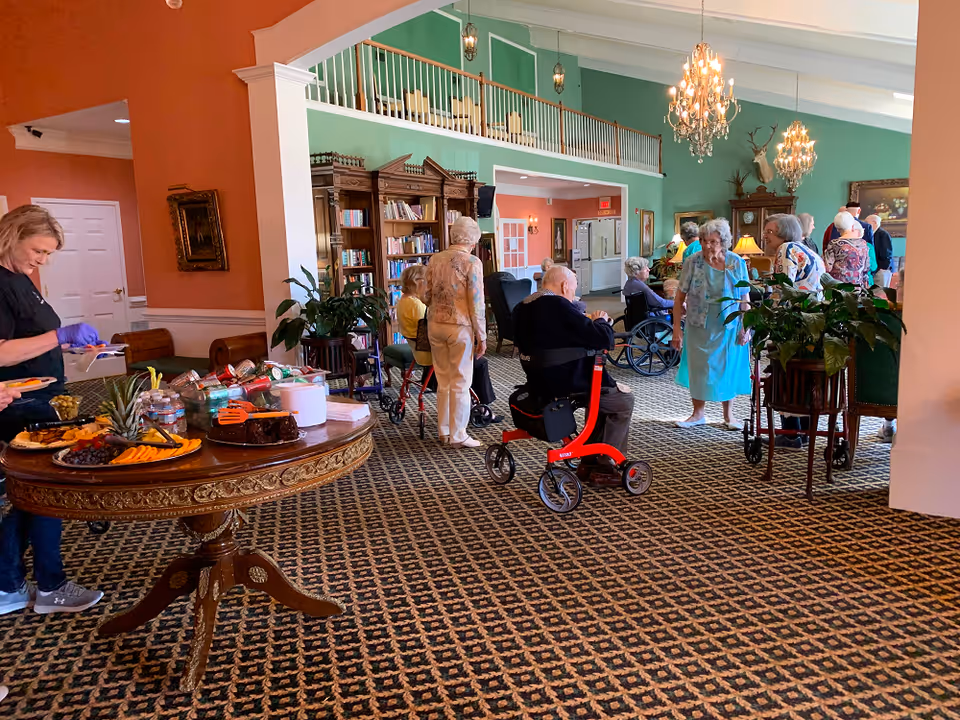 A senior living facility common area with residents gathered around a snack table and seating under chandeliers.