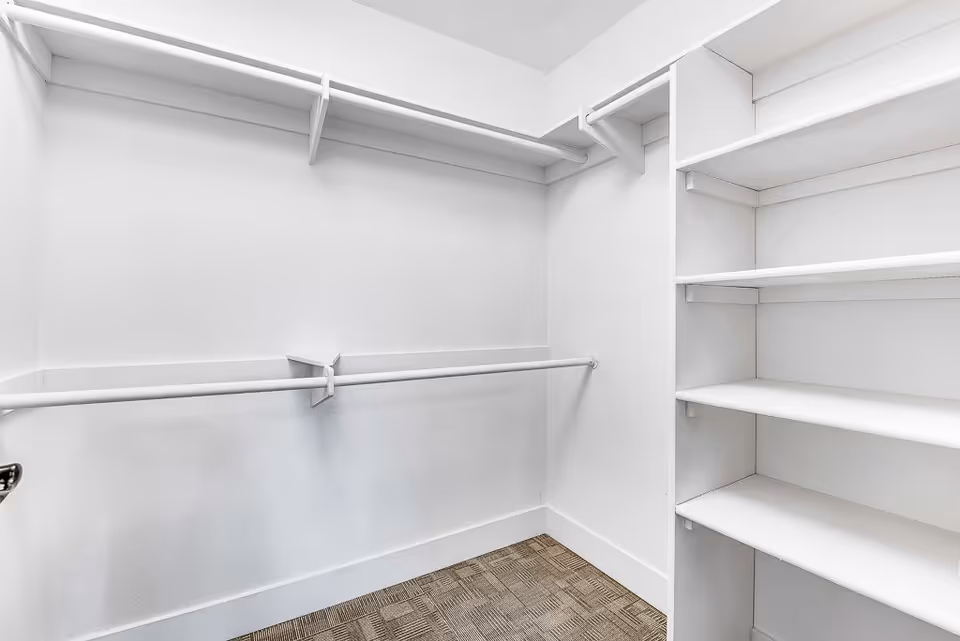 Empty walk-in closet with white walls, multiple hanging rods, and built-in white shelves on the right side. The floor is covered with a patterned carpet.