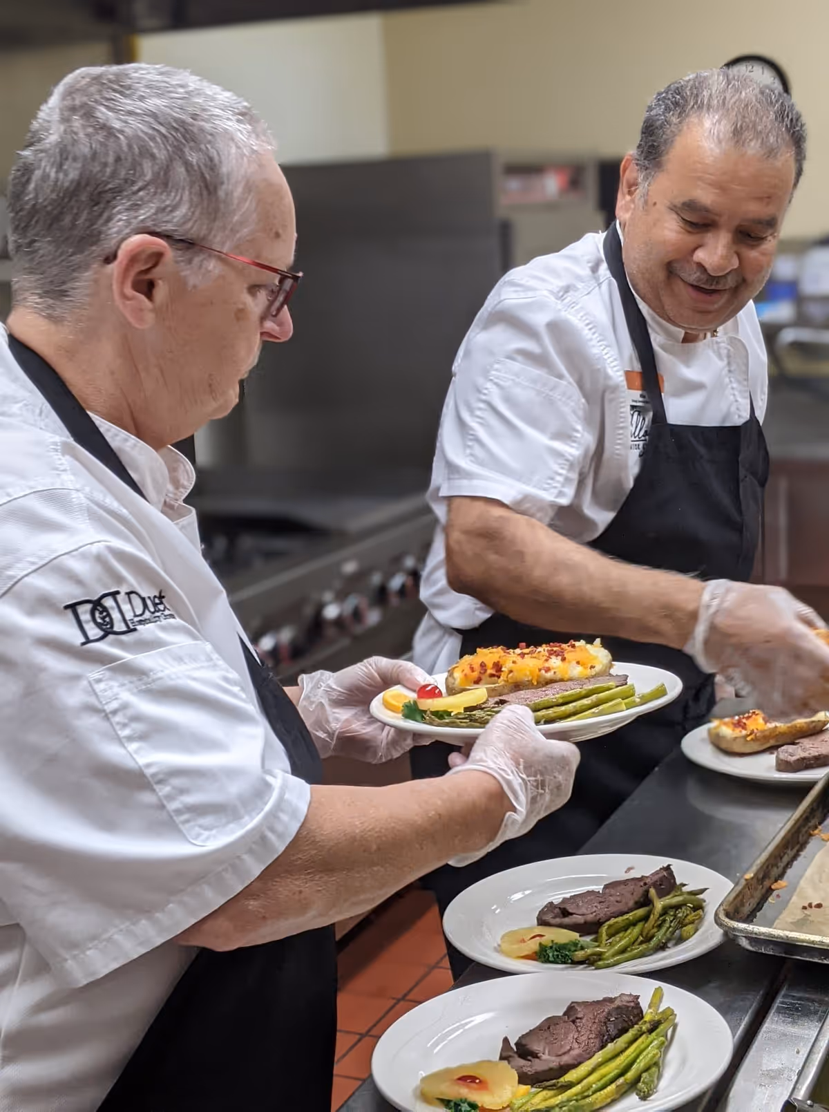 Two chefs in white uniforms and black aprons preparing and plating meals in a commercial kitchen. The plates contain steak, asparagus, pineapple slices, and a twice-baked potato topped with cheese and bacon bits.