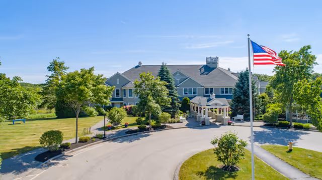 Front entrance of a two-story senior living building with a covered porte-cochère, circular drive, landscaped grounds, and an American flag flying nearby.
