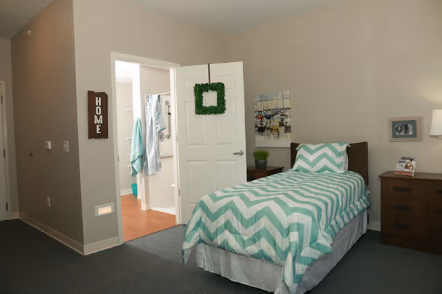 A bedroom with a single bed covered in a green and white chevron patterned bedspread. The bed is positioned against a beige wall with a nightstand beside it holding a lamp, a framed photo, and a small plant. The door to the room is open, revealing a bathroom with towels hanging and a shower curtain visible. A decorative green wreath hangs on the door, and a wooden sign with the word 'HOME' is mounted on the wall near the bathroom entrance.