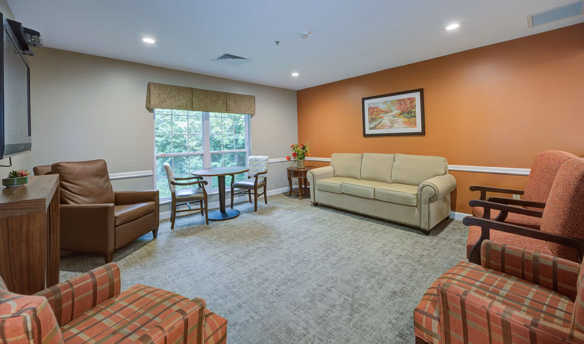 A cozy living room with a beige sofa, two patterned armchairs, a brown leather armchair, a small round table with two chairs near a window, and a wooden TV stand with a mounted TV. The walls are painted beige and orange, with a framed painting hanging above the sofa.