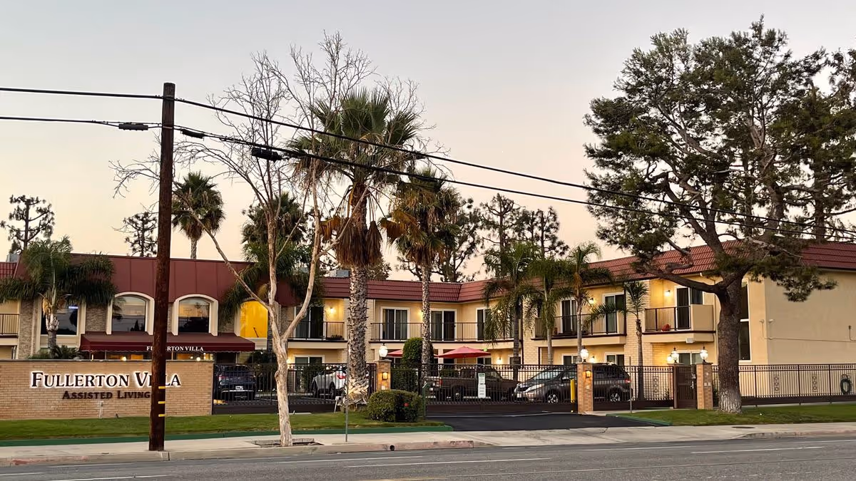 Exterior front of the Fullerton Villa assisted living building with palm trees, a gated entrance, and a street-facing sign at dusk.
