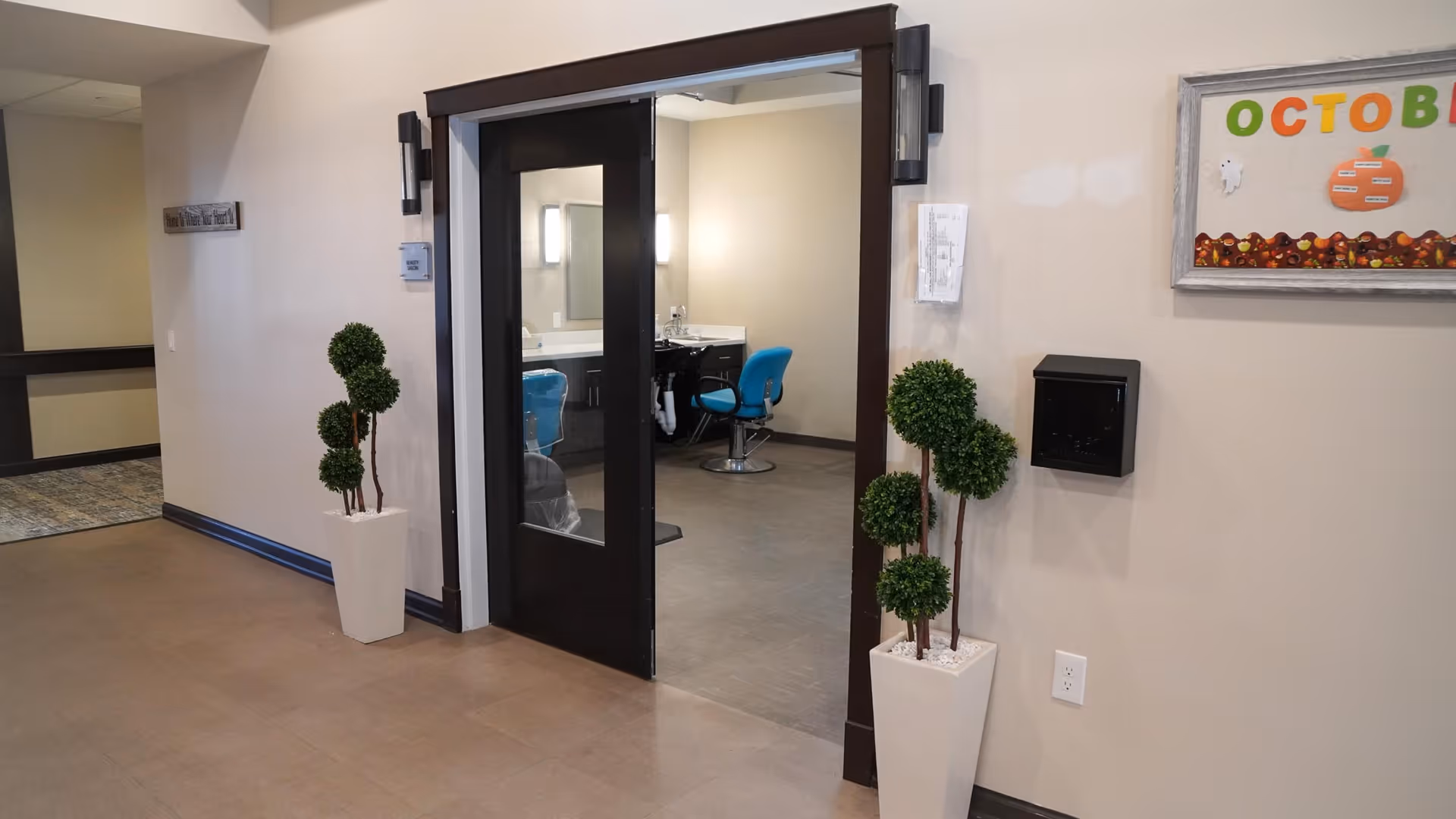 Interior view of a hallway leading to a room with a salon chair and mirror, featuring double dark-framed glass doors, two tall white planters with green topiary plants on either side of the entrance, and a bulletin board on the wall with colorful October decorations.