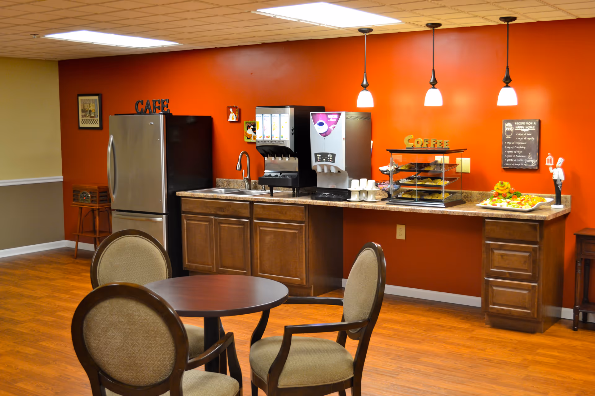 A small cafe area with a round table and four upholstered chairs in the foreground. In the background, there is a countertop with a sink, a stainless steel refrigerator, a beverage dispenser, a coffee station with cups, and a display case with pastries. The wall behind the counter is painted orange and decorated with signs that say 'CAFE' and 'COFFEE', as well as a framed recipe and small decorative items. Three pendant lights hang from the ceiling above the counter.