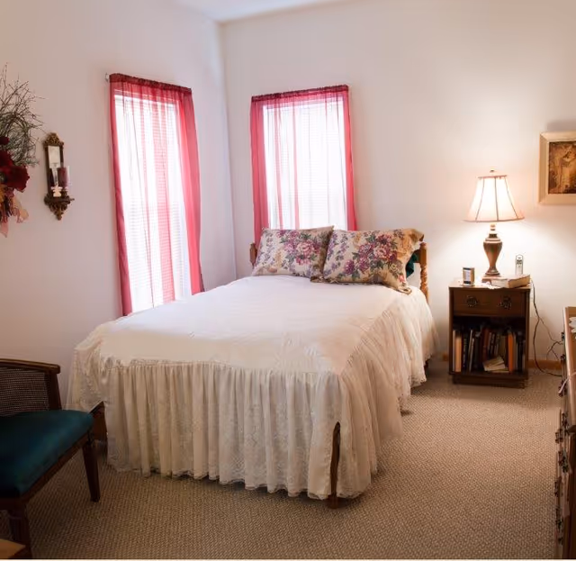 A cozy bedroom with a bed covered in a white bedspread and floral pillows. Two windows with sheer red curtains are behind the bed. A wooden nightstand with a lamp, books, and a phone is beside the bed. A green cushioned chair is partially visible on the left side of the room. The walls are white and the floor is carpeted.