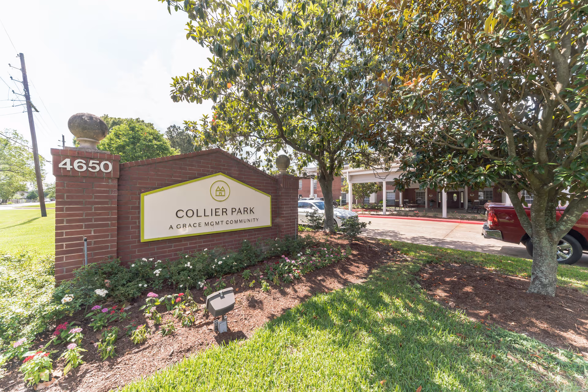 Entrance sign for Collier Park, a Grace Management community, displayed on a brick structure with the address number 4650. The sign is surrounded by landscaped flowers and greenery, with trees and a driveway leading to a building entrance visible in the background.