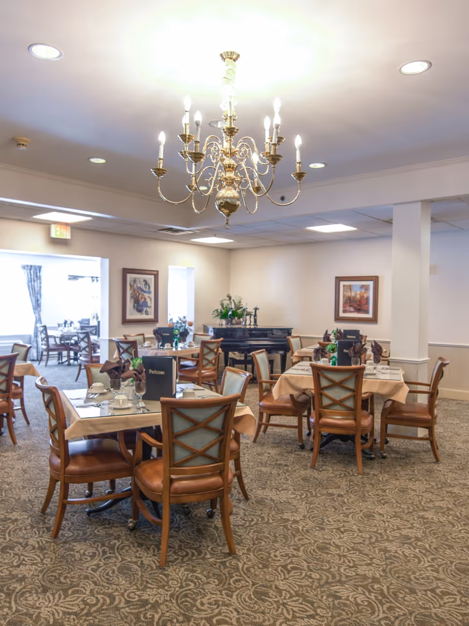 A bright dining room with multiple set tables and wooden chairs beneath a central chandelier.