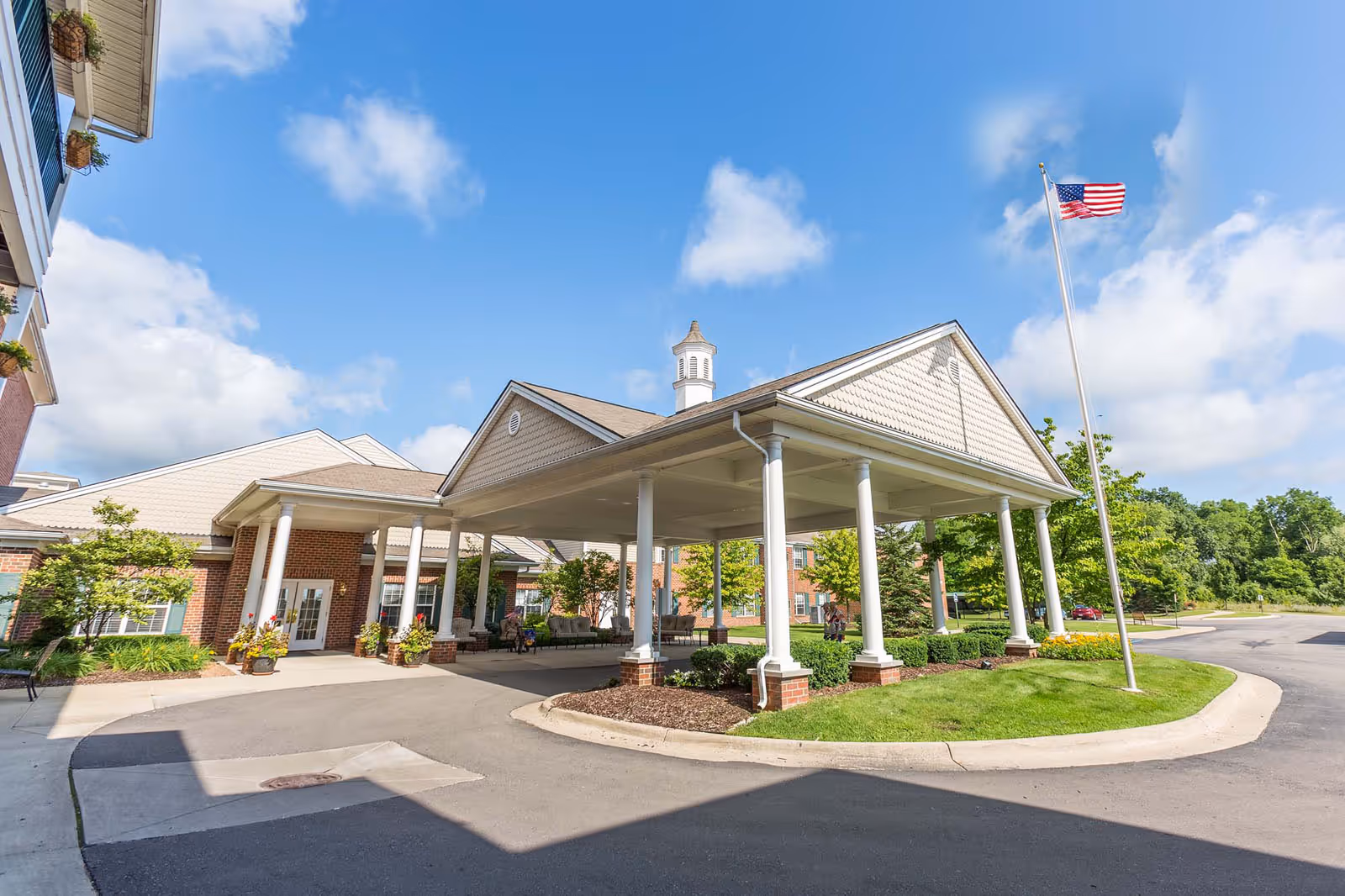 Entrance of a senior living facility with a covered drop-off area supported by white columns, an American flag on a flagpole, and well-maintained landscaping under a bright blue sky with some clouds.
