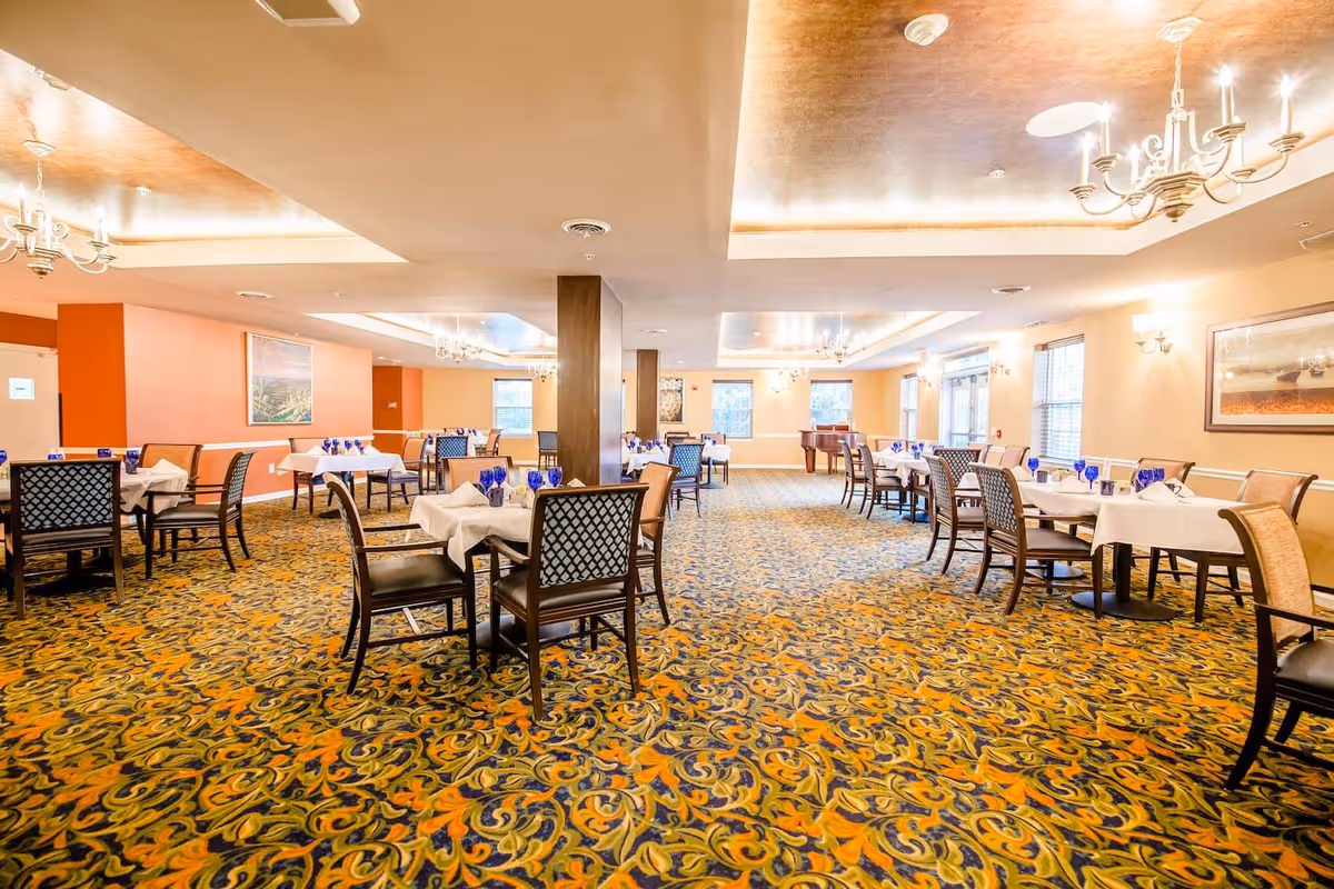A spacious dining room with multiple tables covered in white tablecloths and set with blue glasses and napkins. The room features patterned carpet, chandeliers, large windows, and framed artwork on the walls.