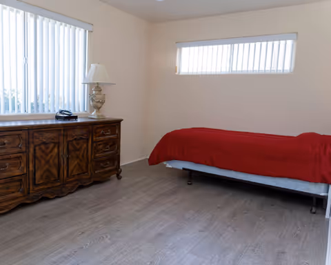 A bedroom with a bed in red bedding, a wooden dresser, and window blinds.