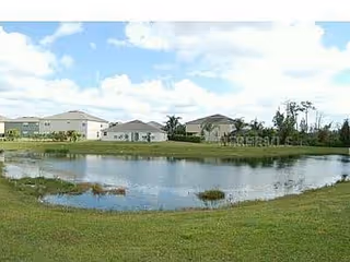 A small pond surrounded by grass with several single-story houses in the background under a partly cloudy sky.