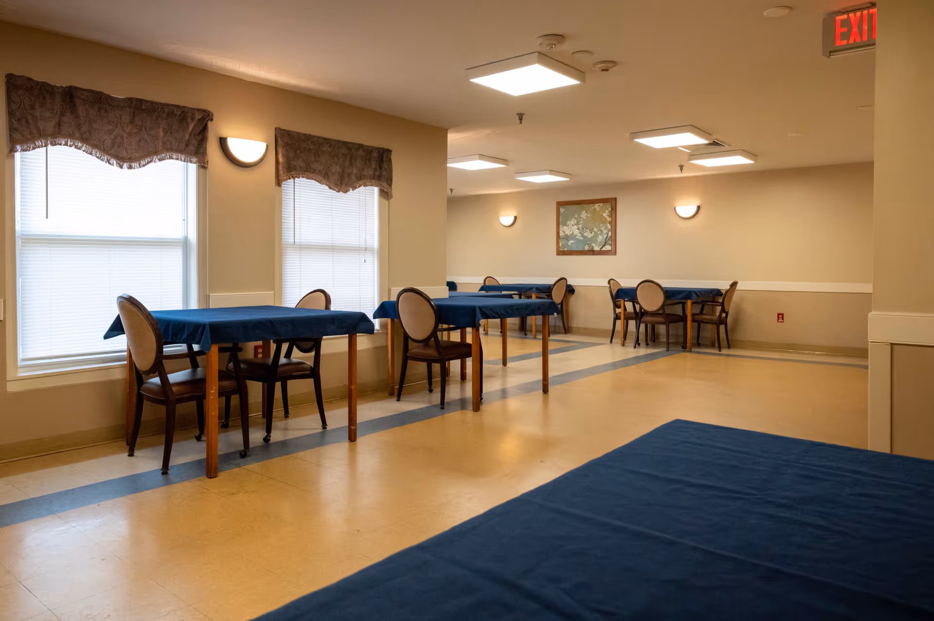 A dining room in a senior living facility with several tables covered in blue tablecloths and wooden chairs with cushioned seats. The room has beige walls, two windows with valances, wall-mounted lights, a framed picture, and an exit sign above a door.