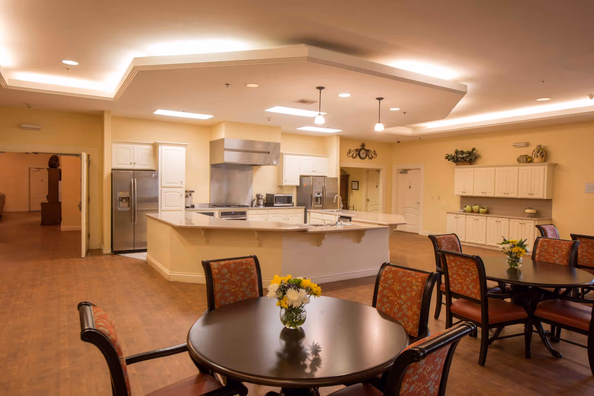 A spacious communal kitchen and dining area with multiple round tables and chairs. The kitchen features white cabinetry, stainless steel appliances including two refrigerators, a stove with a large hood, a microwave, and a central island with a sink. The room has warm lighting and decorative elements such as flowers on the tables and ornamental items on the cabinets.