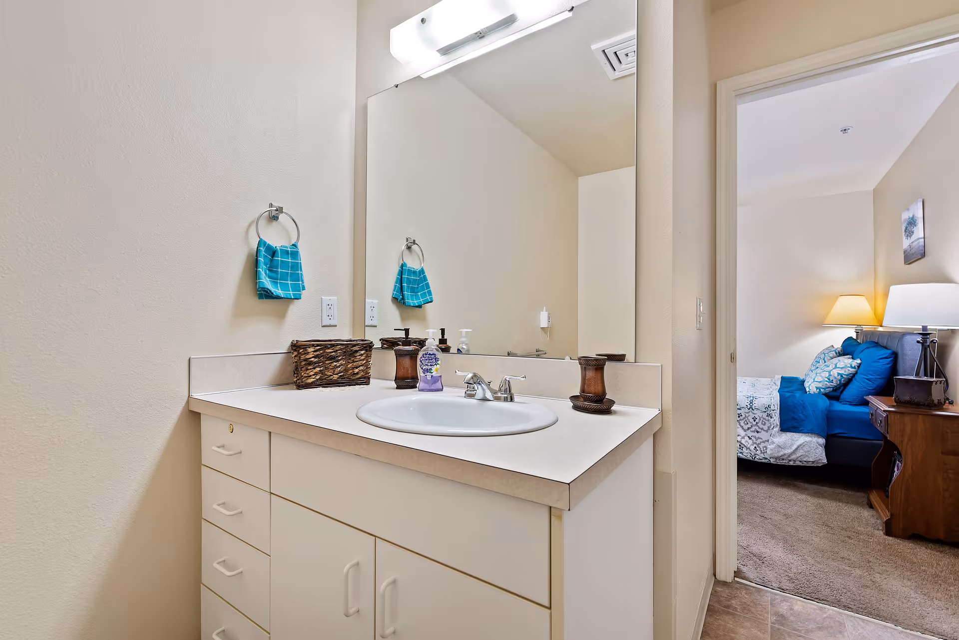 A bathroom vanity with a white countertop, a sink, a large mirror, and a light fixture above. A turquoise hand towel hangs on a ring on the wall. On the countertop, there is a woven basket, a soap dispenser, and a decorative item. Through an open doorway, a bedroom with a bed, blue pillows, a nightstand, and a lamp is visible.
