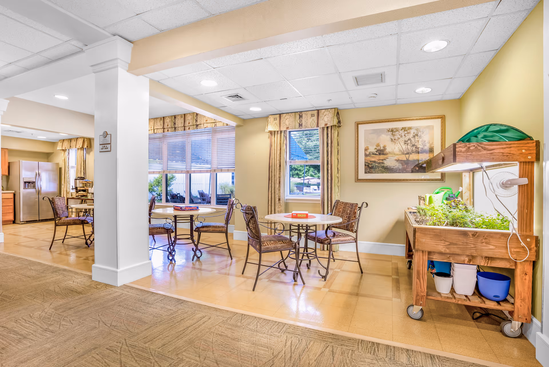 Communal dining area with round tables and chairs, a small kitchenette with a refrigerator, and a wheeled indoor plant/garden cart against a pale yellow wall.