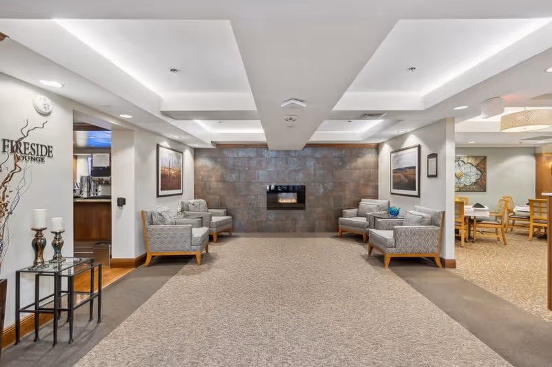 A cozy lounge area with four cushioned armchairs arranged around a modern wall-mounted fireplace. The walls are decorated with framed landscape artwork, and the ceiling features recessed lighting. To the left, a sign reads 'Fireside Lounge' next to a small glass table with decorative candles. To the right, there is a dining area with wooden chairs and tables.
