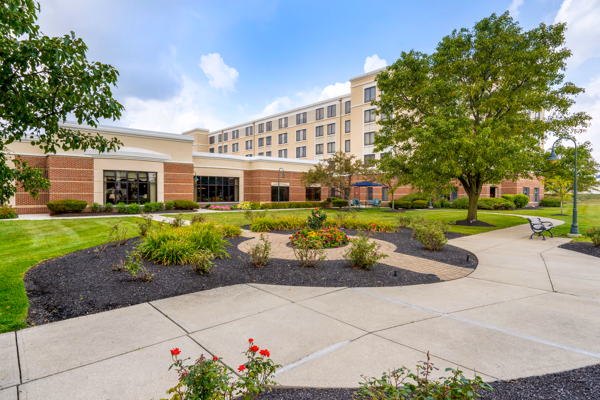 Outdoor garden area with paved walkways, flower beds, bushes, and trees in front of a multi-story senior living facility building under a partly cloudy sky.