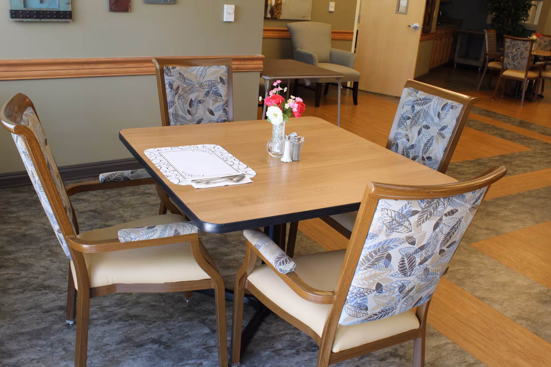 A square wooden dining table set with four upholstered chairs, a placemat, salt and pepper shakers and a small vase of flowers in a communal dining area.