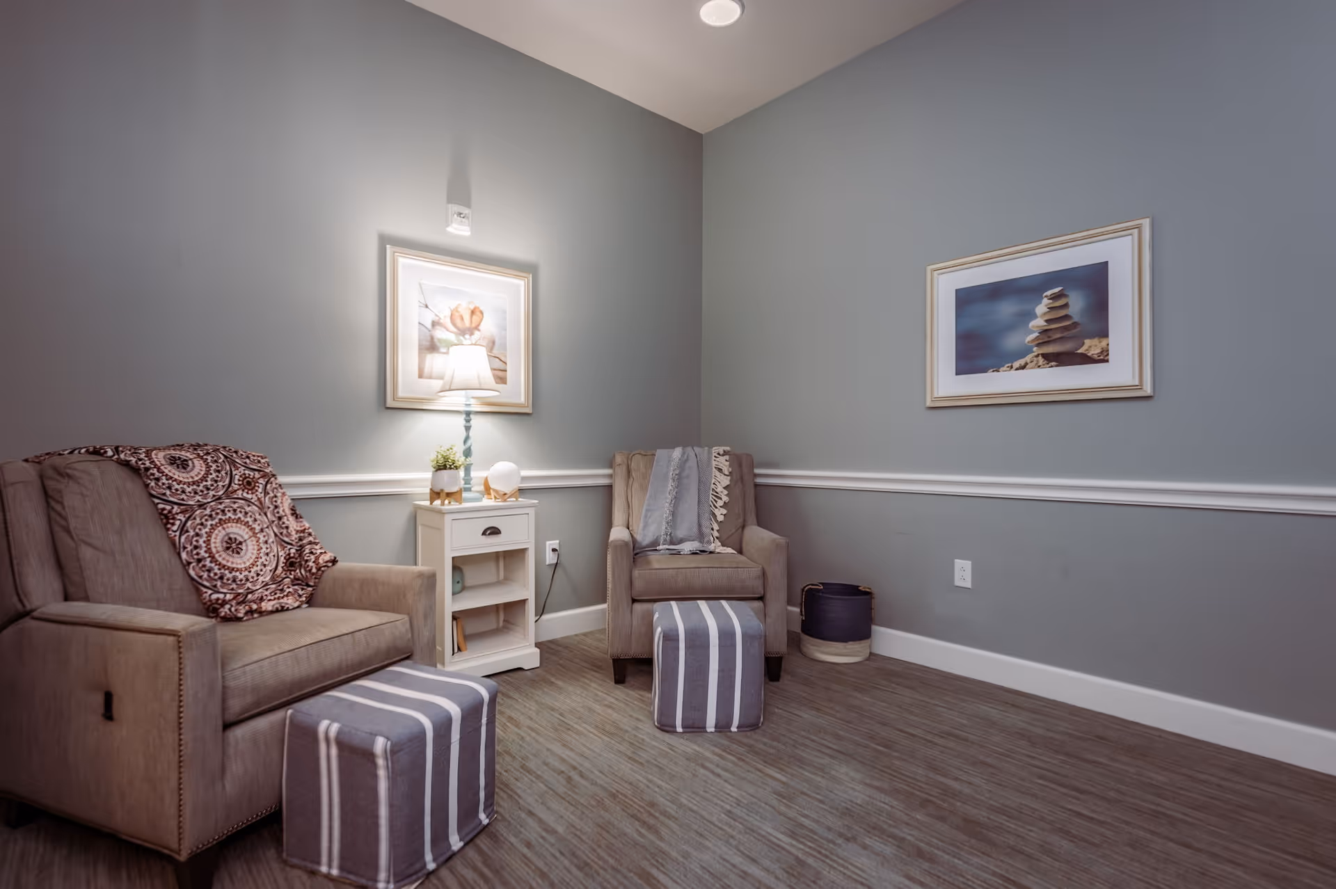 A cozy sitting area with two beige armchairs, each with a striped ottoman in front. A small white side table with a lamp and decorative items is placed between the chairs. The walls are painted gray with white trim, and two framed pictures hang on the walls. The floor has a wood-like finish.
