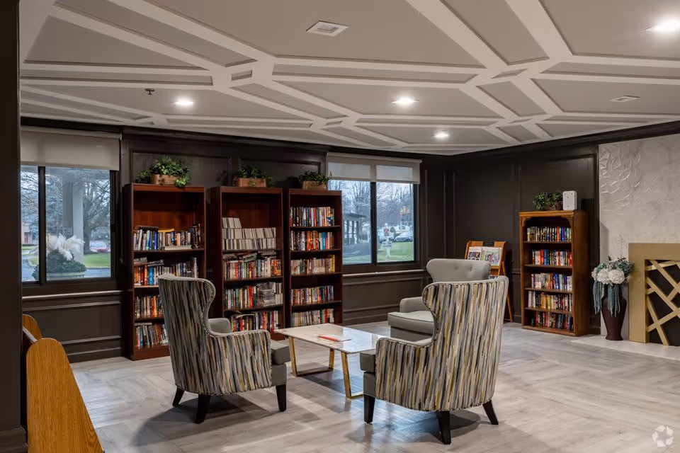 A cozy common room with patterned armchairs around a coffee table and bookshelves against a wall beneath decorative ceiling panels and windows.