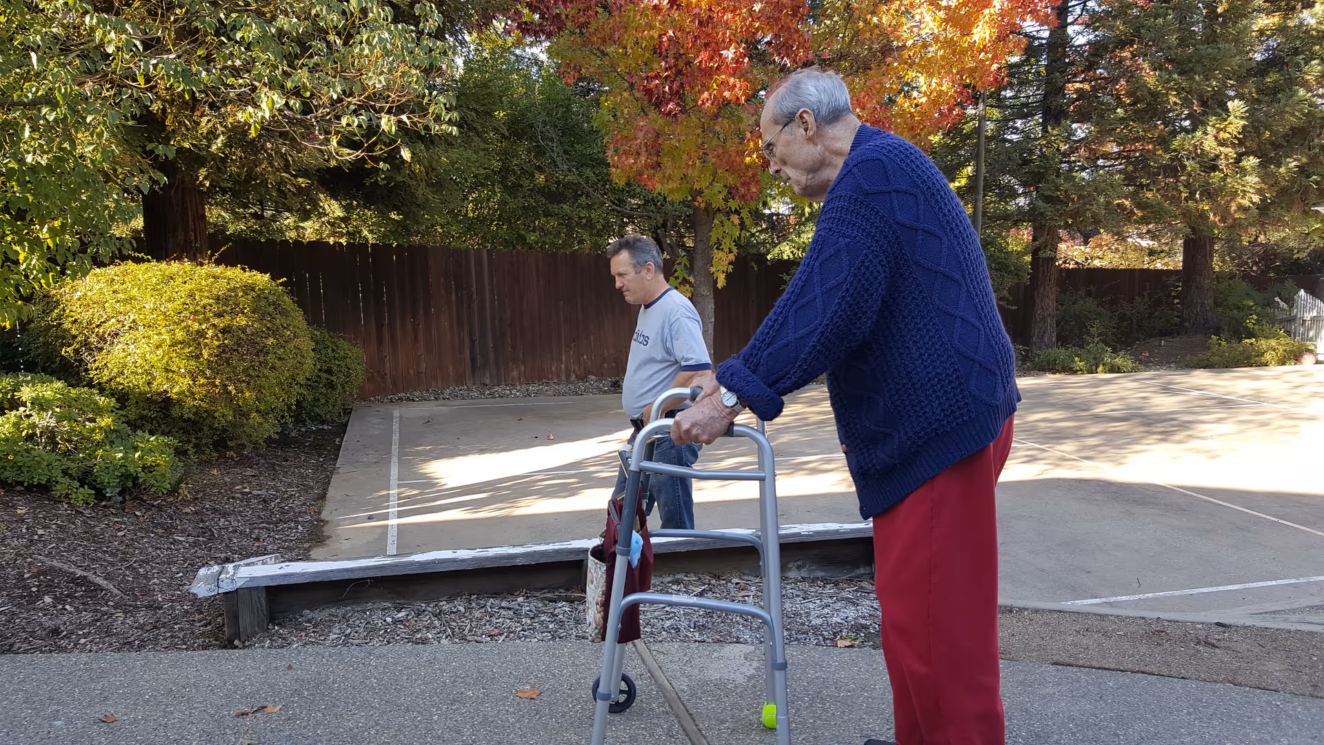 An elderly person using a walker outdoors on a paved area with trees and bushes in the background. Another person is walking nearby. The scene is set in a sunny area with autumn-colored leaves on the trees.