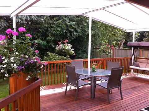 Wooden outdoor deck with table and chairs under a canopy, surrounded by flowering planters.