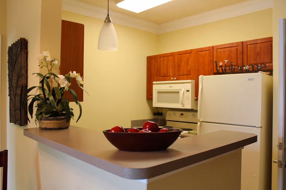 Interior view of a kitchen with wooden cabinets, a white refrigerator, a white microwave above a stove, a countertop with a bowl of red apples, and a potted orchid plant. A pendant light hangs above the counter and a decorative 'Welcome' sign is placed on top of the refrigerator.