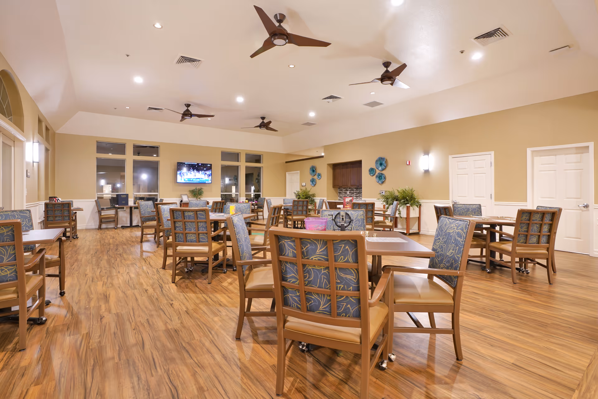 A spacious dining room with multiple wooden tables and chairs featuring patterned blue cushions. The room has a wooden floor, beige walls, ceiling fans, and recessed lighting. A flat-screen TV is mounted on the far wall above a small table with plants and decorative items.
