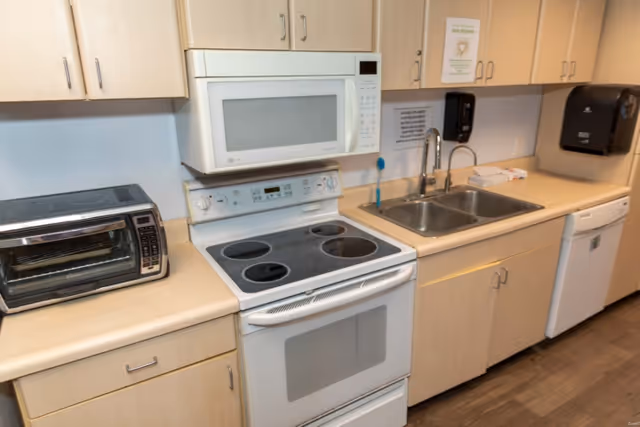 A kitchen area with light wood cabinets, a white electric stove with four burners, a white microwave above the stove, a double stainless steel sink with a faucet, a toaster oven on the counter, and a dishwasher. There are paper towel dispensers and a soap dispenser mounted on the wall above the sink.