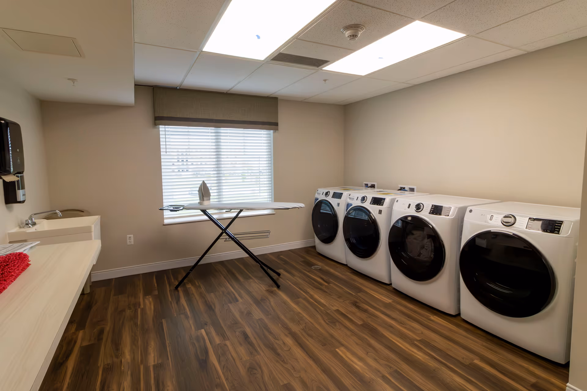 Laundry room with four front-loading washing machines and dryers lined up against a beige wall. An ironing board with an iron is set up in front of a window with blinds. There is a countertop with a sink and a soap dispenser on the left side of the room. The floor is wood-patterned and the ceiling has recessed lighting.