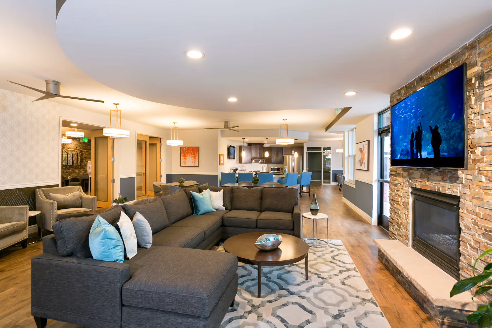 A modern senior living facility common area featuring a large gray sectional sofa with blue and white pillows, a round wooden coffee table, and a stone fireplace with a mounted flat-screen TV. The room has wooden flooring, ceiling lights, and a dining area with blue chairs and a kitchen in the background.