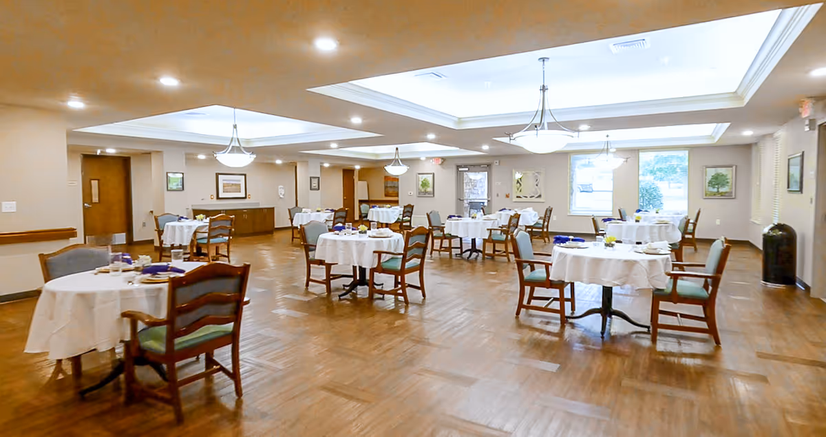 A spacious dining room with multiple round tables covered with white tablecloths, each surrounded by wooden chairs with green cushions. The room has wooden flooring, beige walls, ceiling lights, and large windows letting in natural light. There are framed pictures on the walls and a sideboard against one wall.