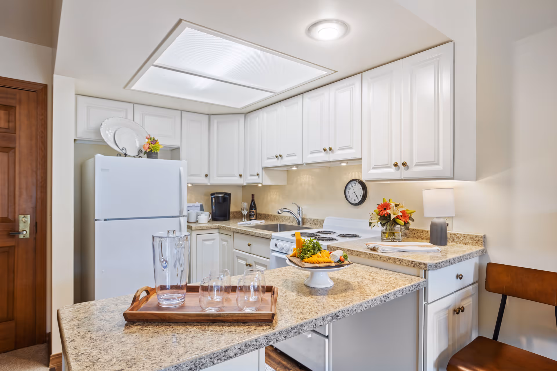 A bright kitchen with white cabinets and granite countertops. There is a white refrigerator, a stove with a clock above it, a coffee maker, and a wooden tray with a glass pitcher and three glasses on the counter. A plate with cheese and vegetables is also on the counter, along with a small lamp and a vase with flowers. A wooden chair is partially visible on the right side.