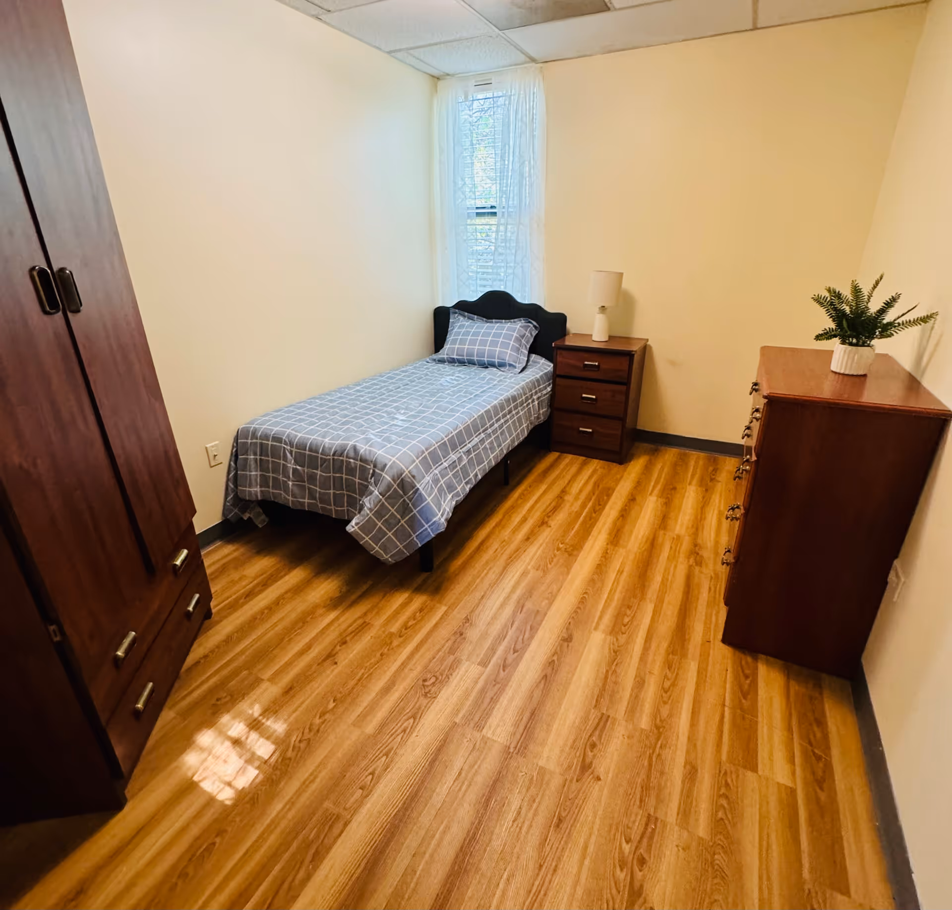 A small, simple bedroom with a single bed covered in a blue and white checkered bedspread. The room has light yellow walls, a wooden floor, a wooden wardrobe on the left, a nightstand with a lamp next to the bed, and a wooden dresser with a small potted plant on top. A window with sheer white curtains is behind the bed.