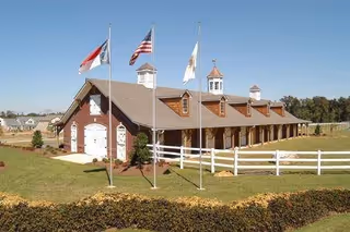 A large barn-style building with a brown roof and white garage doors, surrounded by a white fence and green lawn. Four flagpoles with flags stand in front of the building under a clear blue sky.