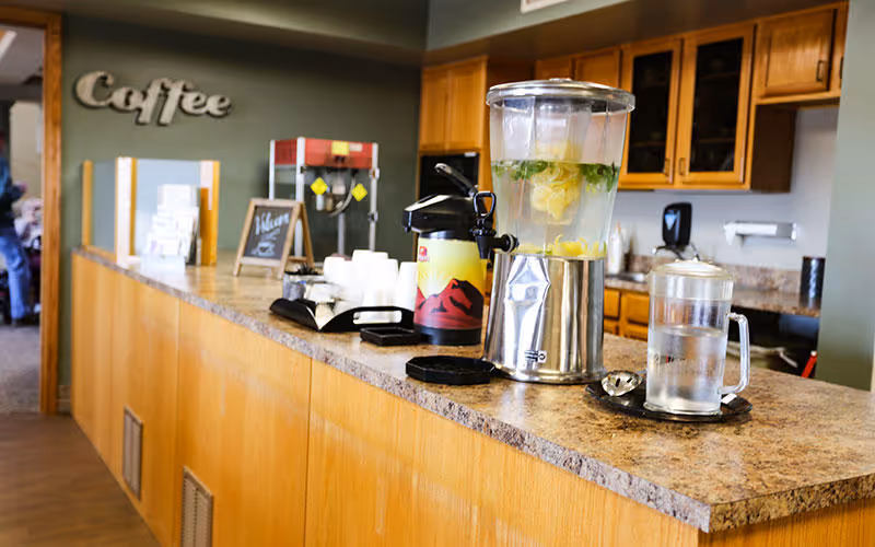 A countertop with a beverage dispenser filled with lemon and mint water, a coffee airpot, disposable cups, and a popcorn machine in the background. The wall behind has wooden cabinets and a sign that says 'Coffee'.