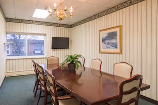 A small conference or meeting room with a polished wooden table surrounded by six upholstered chairs. A potted plant is placed in the center of the table. The room has striped wallpaper with a floral border near the ceiling, a chandelier hanging above the table, a wall-mounted flat screen TV, a framed picture on the wall, and a window showing an outside view of buildings and trees.