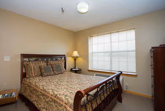 A bedroom with a large window covered by blinds, a wooden bed frame with an ornate headboard and footboard, a bed with patterned bedding and multiple pillows, a bedside table with a lamp, and a wooden dresser in the corner.