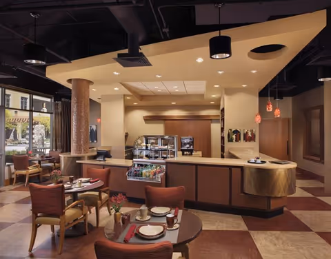 Interior view of a dining area in a senior living facility with round tables set with plates, cups, and napkins. The room features a counter with a display case containing beverages and snacks, pendant lights hanging from the ceiling, and large windows letting in natural light.