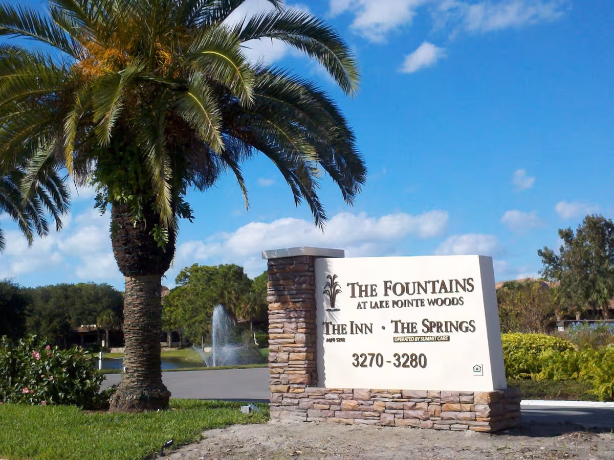 Entrance monument sign for The Fountains at Lake Pointe Woods beside a palm tree with a fountain and landscaped grounds in the background.