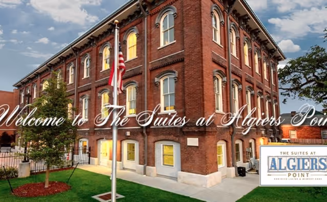 Three-story red brick building with lit windows, an American flag, and landscaping at The Suites at Algiers Point.