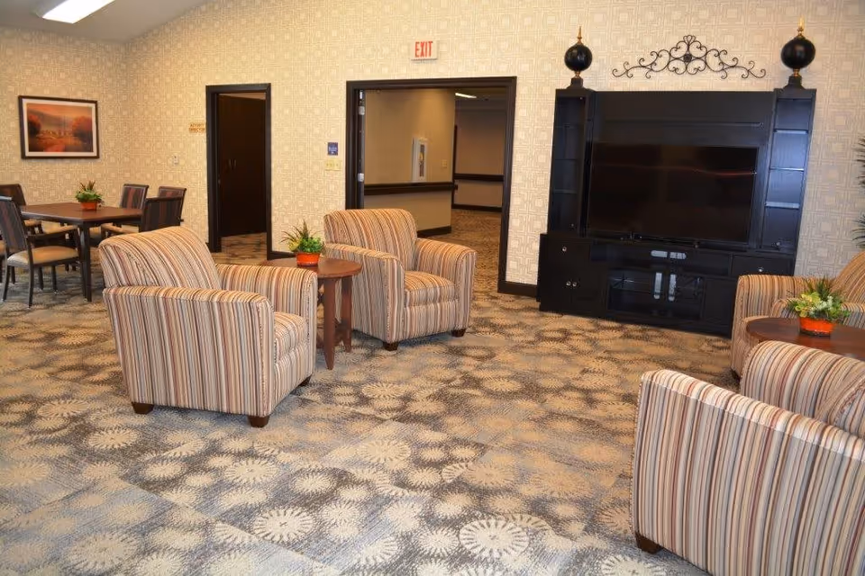 A cozy living room area in a senior living facility featuring four striped armchairs arranged around two small wooden tables with potted plants. A large black entertainment center with a flat-screen TV is against the wall. In the background, there is a dining area with a table and chairs, and two open doorways leading to hallways. The room has patterned carpet and light-colored wallpaper with a geometric design.