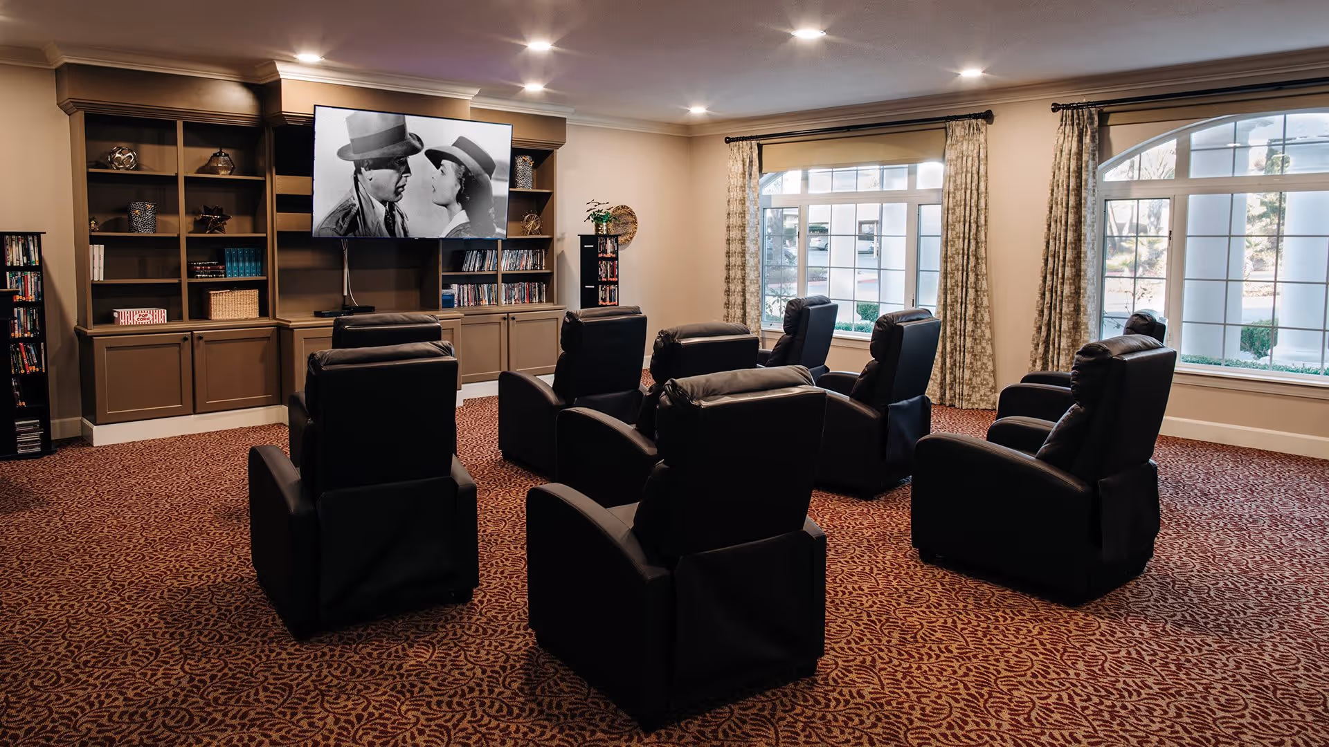 A cozy media room with eight black recliner chairs arranged in two rows facing a large flat-screen TV mounted on a built-in wooden shelving unit. The TV is displaying a black and white movie scene of two people wearing hats. The room has patterned carpet, large windows with curtains, and soft ceiling lighting.