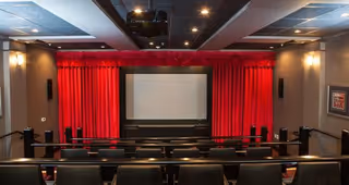 Interior view of a small theater room with rows of black seats facing a large screen framed by red curtains. The room has dim lighting with ceiling lights and speakers mounted on the walls.