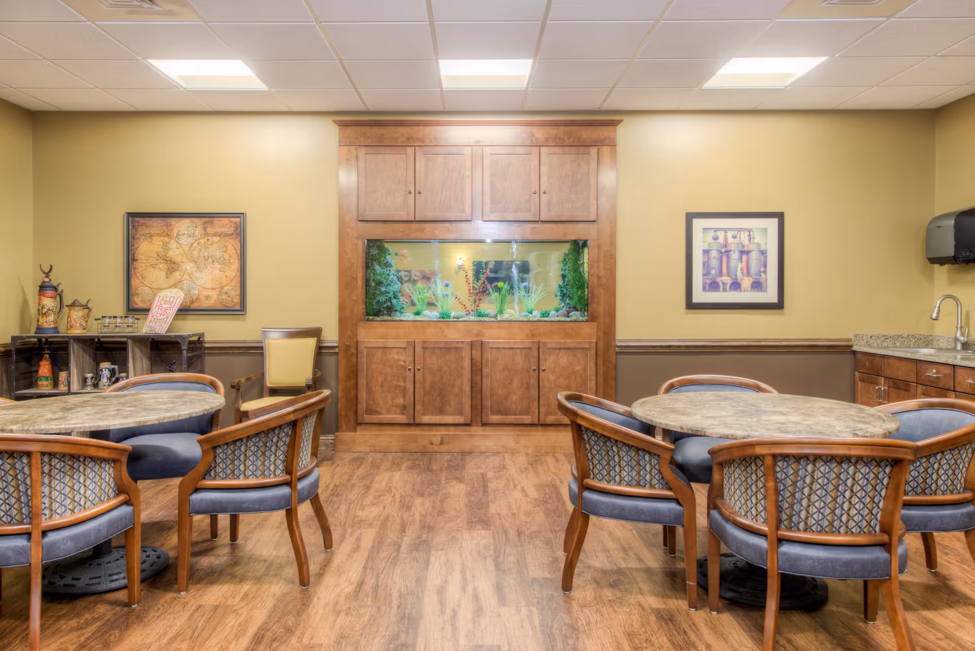 A cozy common area with two round tables surrounded by wooden chairs with blue cushions. In the center of the back wall is a built-in wooden cabinet with an aquarium filled with plants and fish. The room has wood flooring, beige walls, framed artwork, and a small kitchenette with a sink and paper towel dispenser on the right side.