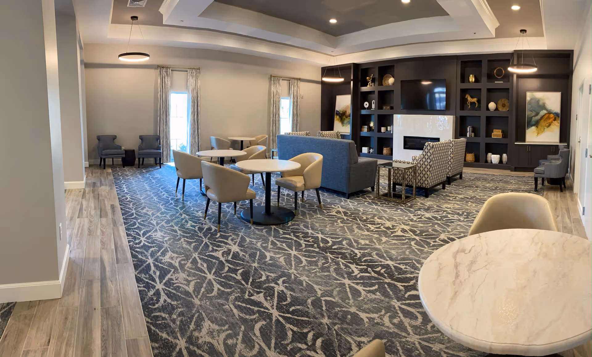 A spacious and well-lit common area with patterned carpet and wood flooring. The room features several round tables with beige chairs, a seating area with sofas and armchairs facing a large built-in dark wood entertainment center with a TV and decorative items. Two large windows with curtains allow natural light to enter the room.